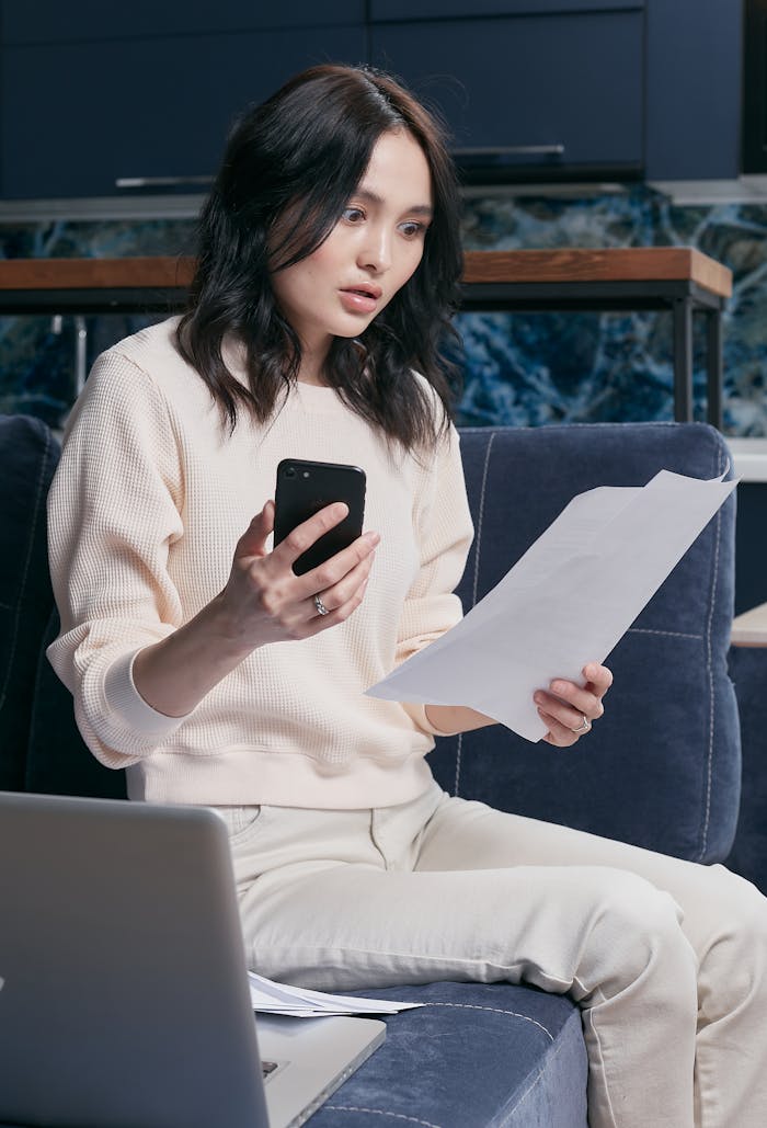 Services-02 A young woman uses her smartphone and laptop to manage finances, appearing focused and concerned.